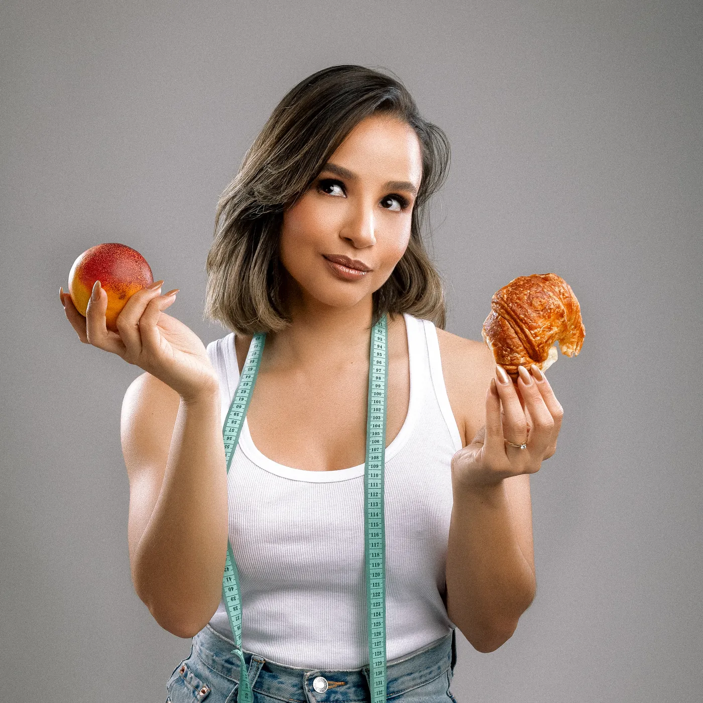 Mairim con una manzana en una mano y un croissant en la otra, con una cinta de medir al cuello